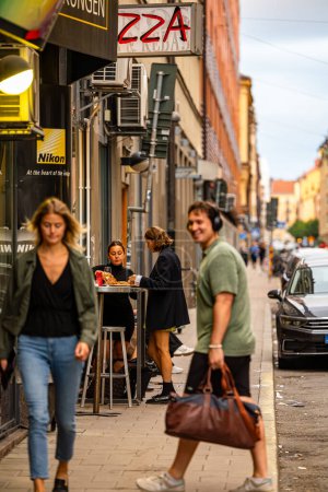 Foto de Estocolmo, Suecia Sept 5, 2023 Dos mujeres jóvenes en la calle en el distrito de Sodermalm comiendo pizza mientras un hombre y una mujer pasan caminando. - Imagen libre de derechos