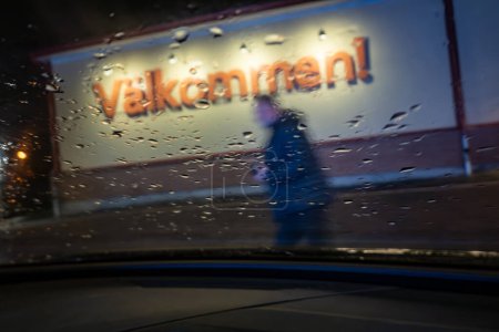 Foto de Varnamo, Suecia Dic 21, 2023 Un hombre pasa por el parabrisas de un coche estacionado y un cartel dice Welcome in Swedish. - Imagen libre de derechos