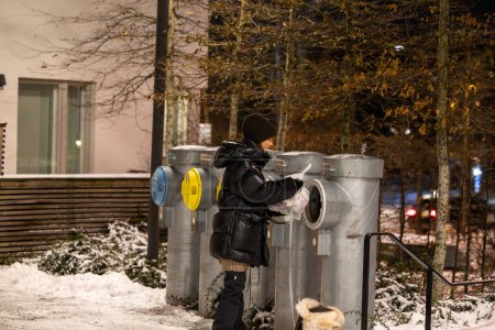 Photo for Stockholm, Sweden, A young woman throws garbage in a shute with a dog on a leash in a resdiential complex at night in the winter. - Royalty Free Image