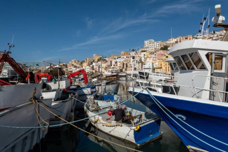 Photo for Sciacca, Sicily, Italy A fishing boat crew member works on a dingy between the fishing boats in the old fishing port and city skyline. - Royalty Free Image