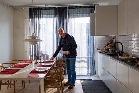 A middle aged man sets the table in a modern stylish kitchen filled withsunlight.