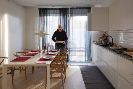 Ein Mann mittleren Alters deckt den Tisch in einer modernen, stilvollen Küche mit Sonnenlicht.