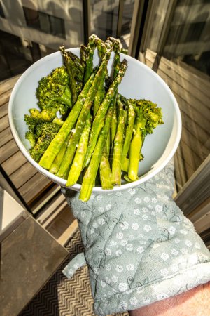 A hand holds cooked asparagus in a bowl with broccoli.