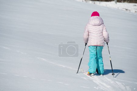 Téléchargez les photos : Vue arrière d'une jeune fille traversant une neige fraîche avec des skis de fond. Enfant jouissant de la liberté hivernale. - en image libre de droit
