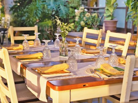 Téléchargez les photos : Une table rectangulaire en bois rustique avec assiettes et verres et serviettes jaunes et six chaises. La table est à l'extérieur dans le jardin, éclairée par le soleil doré du soir. Plantes vertes luxuriantes en arrière-plan. Pas de peuple. - en image libre de droit