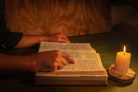 Photo for Hands of a man studying the bible pointing to a bible passage by candlelight on a table - Royalty Free Image
