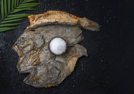 Téléchargez les photos : Open salted fish on a black textured background with a well of sea salt and a palm leaf - en image libre de droit