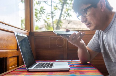 Téléchargez les photos : Homme mûr envoyant un message audio d'un chalet à côté de son ordinateur portable. Photo de haute qualité - en image libre de droit
