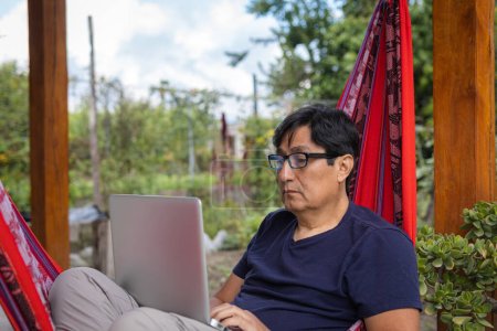 Téléchargez les photos : Gros plan d'un homme hispanique avec des lunettes travaillant à distance sur un ordinateur portable à partir d'un hamac. Photo de haute qualité - en image libre de droit
