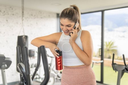 Téléchargez les photos : Souriant jeune femme parlant sur téléphone portable avec bouteille d'eau réutilisable dans une salle de gym le matin - en image libre de droit