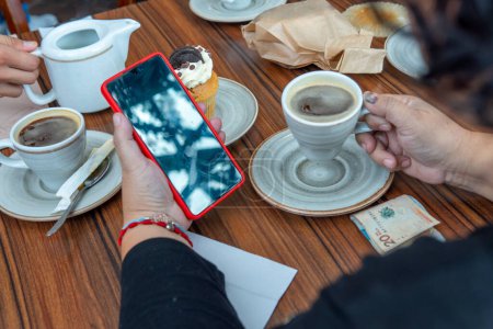 Unerkennbare Frau trinkt eine Tasse Kaffee, während sie in einer Cafeteria neben einem Cupcake und einem 20-kolumbianischen Peso-Schein auf ihr Handy blickt