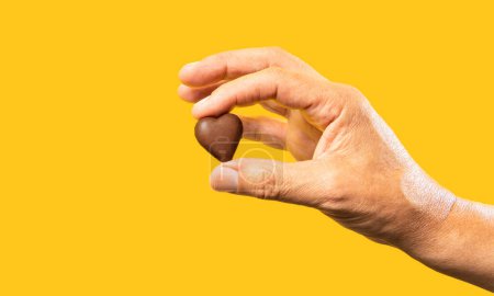 Die Hand eines unkenntlich gemachten Mannes auf gelbem Hintergrund hält eine herzförmige Schokolade. Konzept der Schokoladenliebe