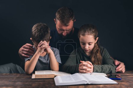 Dad and children are praying. An open Bible on the table. A group of Christians are praying. In the room