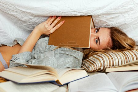 Cute blonde woman in white shirt on the bed in home bedroom, reads a book. Model under a blanket cover with a book. Many books in the foreground
