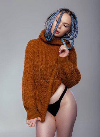 Attractive model with black panties and turtleneck warm sweater posing provocatively. Slim beautiful woman with long pigtails hairstyle, sporty figure is posing on a gray background.