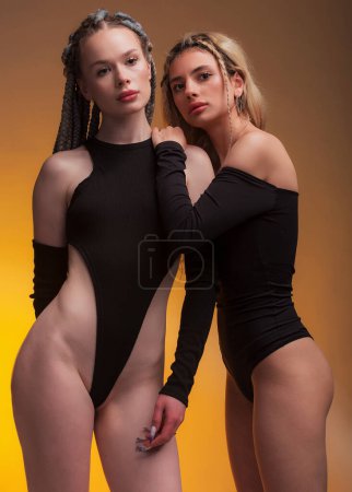 Photo for Two girlfriend hugging each other, feeling love, posing in black bodysuit with long hair, perfect lips and skin, looking in camera on warm orange background. - Royalty Free Image