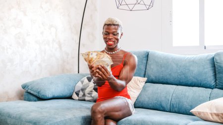 Photo for Excited African American person in stylish red dress with makeup examining bunch of money while sitting on sofa in living room at home - Royalty Free Image