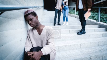 Upset African American transgender student leaning on wall near crop classmates throwing crumpled paper outside university building