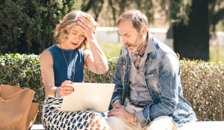 Seniorpaar schaut traurig und enttäuscht, während es einen Laptop in einem sonnigen Park benutzt, was darauf hindeutet, dass es online Geld verloren haben könnte