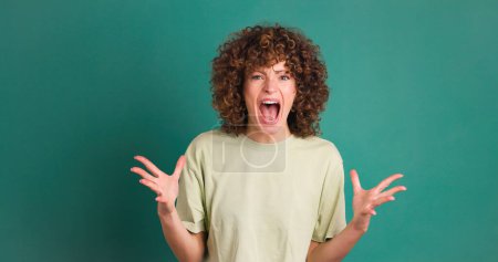 Téléchargez les photos : Jeune femme aux cheveux bouclés regardant la caméra tout en se tenant debout en hurlant avec les mains tendues secouant la tête et les doigts ouverts sur fond vert - en image libre de droit
