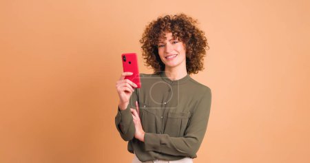 Photo for Smiling young woman in pink outfit with curly hair browsing smartphone while pointing away on beige background - Royalty Free Image