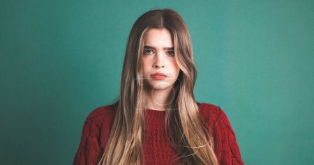 Modelo femenina joven deprimida en suéter granate con pelo largo y rubio mirando a la cámara sobre fondo verde
