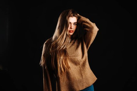 Retrato de sensual modelo femenino joven en suéter marrón posando con la mano en pelo largo y rubio mirando a la cámara mientras está de pie sobre fondo negro