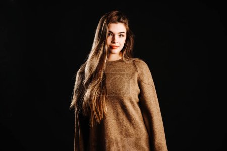 Retrato de hermosa modelo femenina joven en suéter marrón con pelo largo y rubio mirando a la cámara mientras está de pie sobre fondo negro