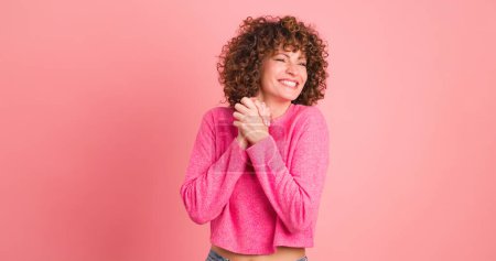 Astonished young curly haired female in makeup and opened mouth looking at camera while standing with folded hands and touching hair on pink background
