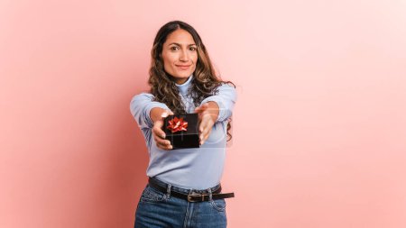 Smiling young Colombian female model wearing casual clothes looking at camera and standing with gift box decorated with bow against pink background in studio