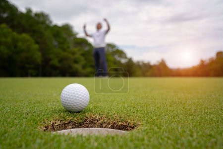 Biała piłka golfowa toczenia w dół otwór golfowy na wprowadzenie zieleni z wieczornym tle pola golfowego i niewyraźne golfistów świętujących w zwycięskim turnieju.