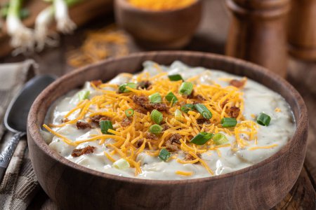 Photo for Creamy potato soup with bacon pieces, shredded cheddar cheese and chopped green onions in a wooden bowl - Royalty Free Image