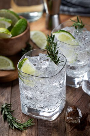 Téléchargez les photos : Gin and tonic cocktail with lime slices, rosemary and ice on a rustic wooden table - en image libre de droit