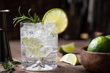 Foto de Gin and tonic cocktail with lime slices, rosemary and ice on a rustic wooden table. Low angle view and copy space - Imagen libre de derechos
