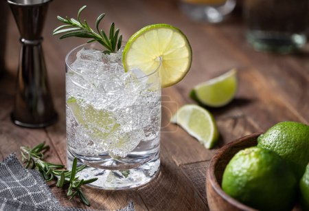 Foto de Gin and tonic cocktail with lime slices, rosemary and ice on a rustic wooden table - Imagen libre de derechos