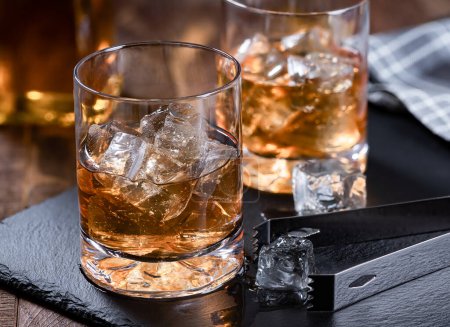 Foto de Dos vasos de whisky con hielo sobre una pizarra negra con fondo de madera - Imagen libre de derechos