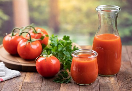 Foto de Jugo de tomate en vaso y jarra con tomates frescos y perejil sobre mesa de madera con fondo de verano - Imagen libre de derechos
