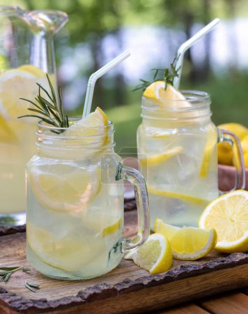 Téléchargez les photos : Citronnade dans deux bocaux en verre avec tranches de citron et romarin sur la table à l'extérieur avec fond de nature - en image libre de droit