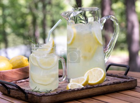 Téléchargez les photos : Verre et pichet de limonade avec tranches de citron et romarin sur plateau en bois avec fond nature extérieur - en image libre de droit