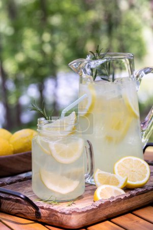 Téléchargez les photos : Verre et pichet de limonade avec tranches de citron et romarin sur plateau en bois avec fond nature extérieur - en image libre de droit