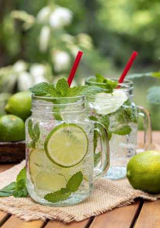 Téléchargez les photos : Cocktail mojito avec tranches de citron vert et menthe à l'extérieur sur table en bois avec fond nature - en image libre de droit