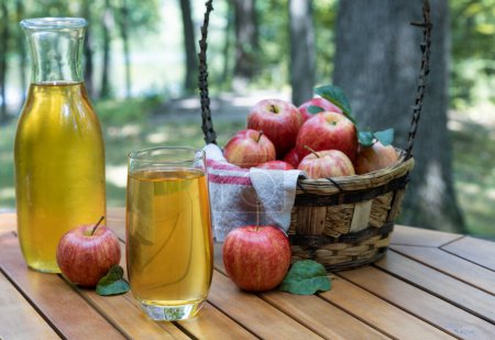Photo for Apple juice in glass and carafe with fresh red apples outdoors on wooden patio table and nature background - Royalty Free Image