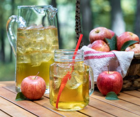 Photo for Apple juice in glass jar and pitcher with fressh red apples outdoors on wooden patio table and nature background - Royalty Free Image