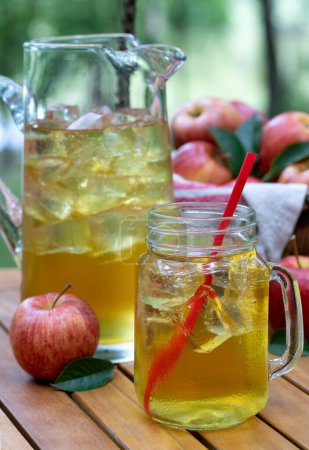 Téléchargez les photos : Jus de pomme dans un bocal en verre et pichet avec pommes rouges fressh à l'extérieur sur une table de patio en bois et fond de nature - en image libre de droit