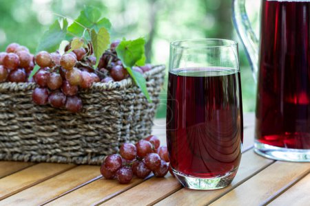 Zumo de uva en vaso y jarra con uvas rojas en una canasta sobre mesa de patio de madera con fondo natural