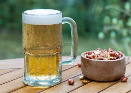 Foto de Taza de vidrio de cerveza y tazón de nueces al aire libre en la mesa del patio de madera con fondo natural - Imagen libre de derechos