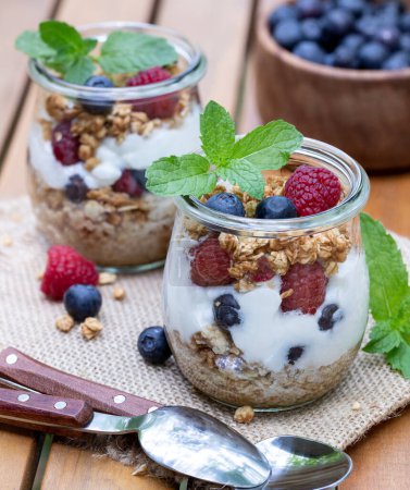 Foto de Parfait de yogur con granola, arándanos y frambuesas decorado con menta al aire libre en la mesa del patio de madera - Imagen libre de derechos