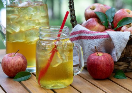 Téléchargez les photos : Jus de pomme dans un bocal en verre et pichet avec pommes rouges fressh à l'extérieur sur une table de patio en bois et fond de nature - en image libre de droit