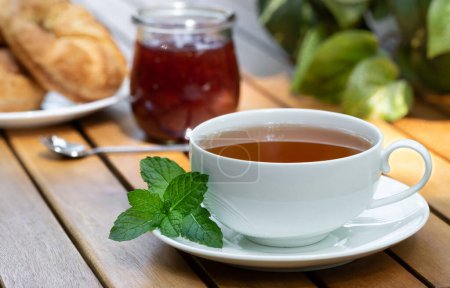 Foto de Taza de té con hojas de menta fresca y croissants y tarro de mermelada en el fondo en la mesa del patio de madera - Imagen libre de derechos