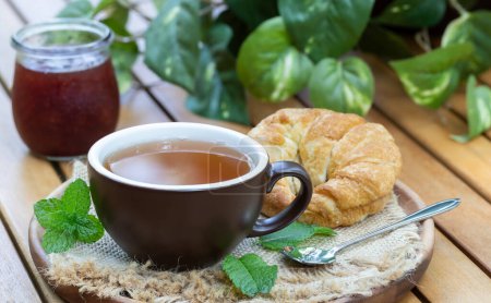 Foto de Taza de té con hojas de menta, croissant y tarro de mermelada al aire libre en una mesa de madera patio - Imagen libre de derechos